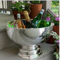 The Hôtel Silver Round Pedestal Chiller sits on a porch step, filled with wine, champagne, and sparkling water bottles amidst potted flowers—ideal for stylish outdoor entertaining with silver-plated elegance.