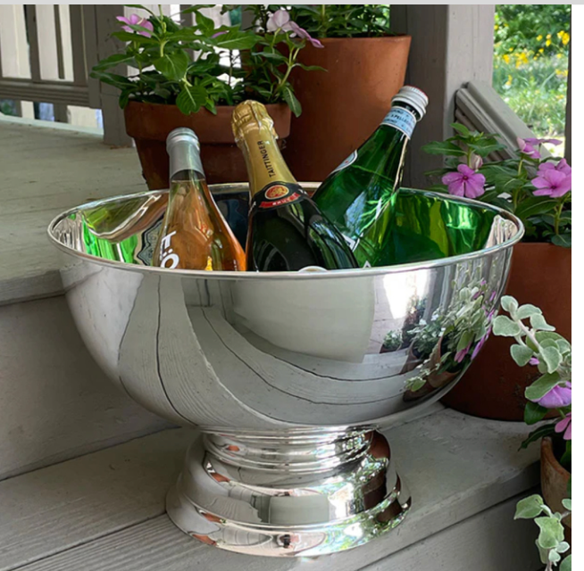 The Hôtel Silver Round Pedestal Chiller sits on a porch step, filled with wine, champagne, and sparkling water bottles amidst potted flowers—ideal for stylish outdoor entertaining with silver-plated elegance.