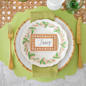 A table setting with green and gold accents, featuring a leaf-patterned plate, gold utensils, a green napkin, and a Hester & Cook Rattan Place Card labeled "Tracy.