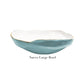 A large ceramic bowl from Beatriz Ball's Encanto Turquoise Serveware Collection featuring a wavy rim, blue exterior, white interior, and thin gold edge. "Nuevo Large Bowl" is displayed beneath the bowl.