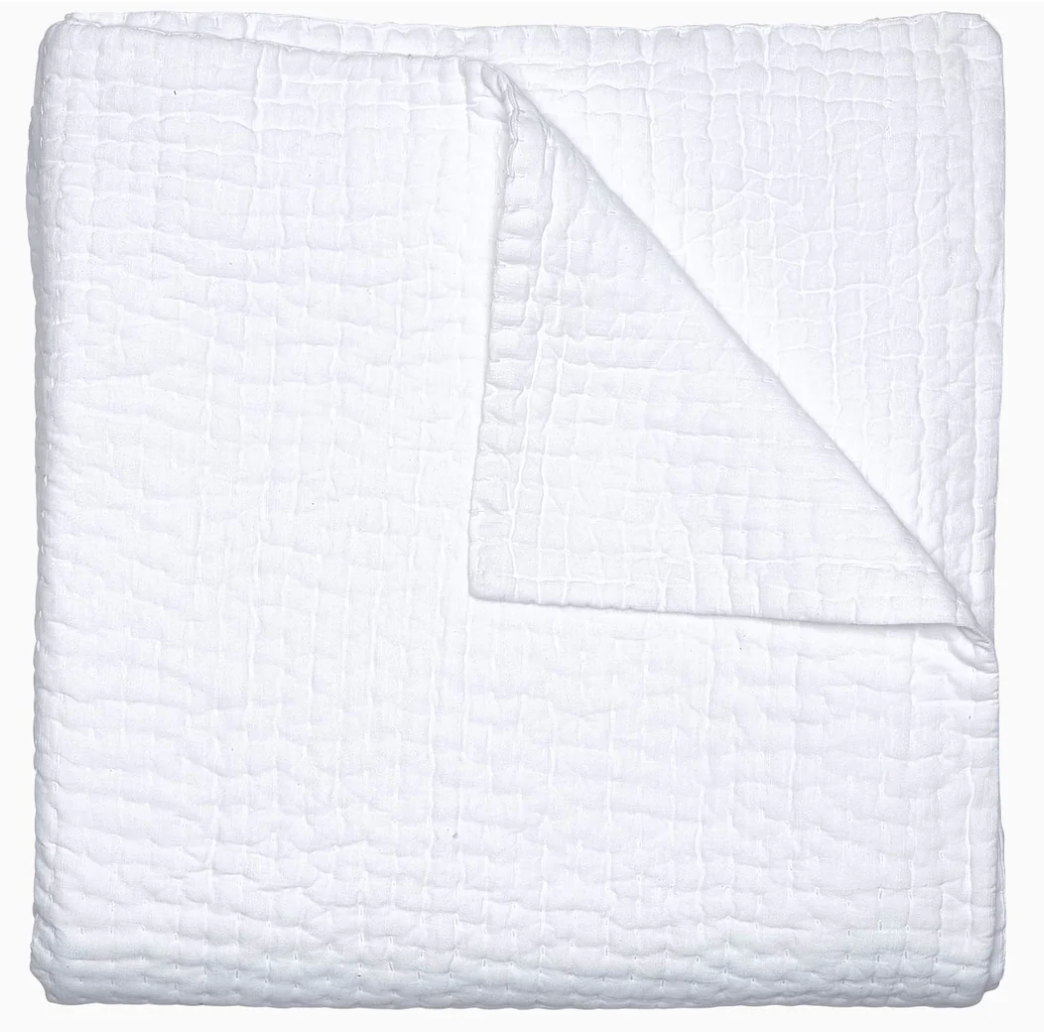 A neatly folded white chambray quilt from the Vivada Bedding Collection by John Robshaw features a textured, grid-like pattern; one corner is turned over to showcase its reversible design and the fine hand-stitched detailing on the underside.