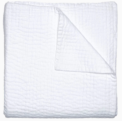 A neatly folded white chambray quilt from the Vivada Bedding Collection by John Robshaw features a textured, grid-like pattern; one corner is turned over to showcase its reversible design and the fine hand-stitched detailing on the underside.