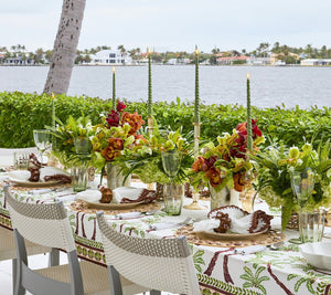 Experience an elegant outdoor dining setup by the waterfront with an island vibe, featuring floral centerpieces and green candles atop the Kim Seybert Oasis Tablecloth. Surrounded by white chairs, swaying palms and houses add to the picturesque view across the water.