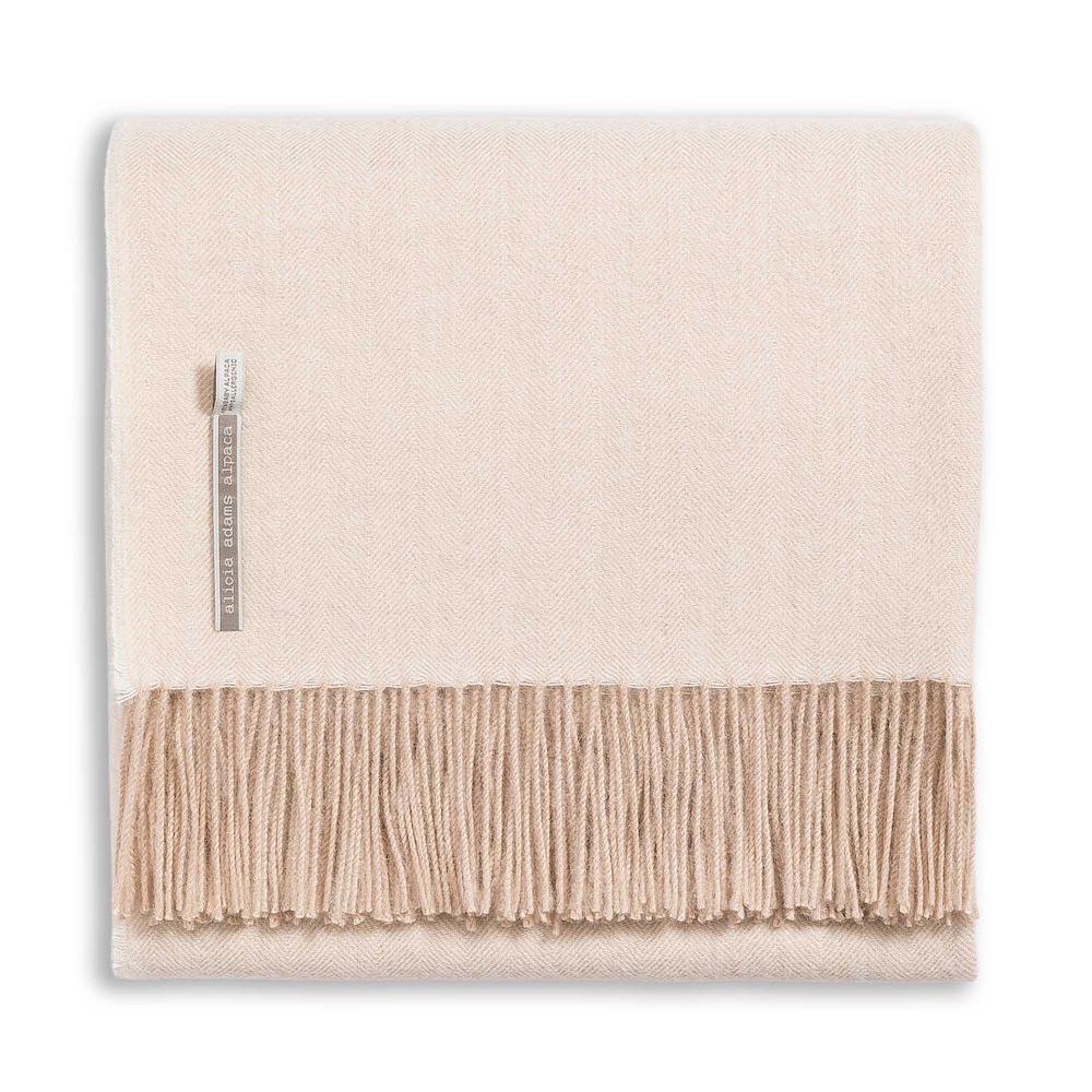 The Alicia Adams Alpaca Classic Herringbone Throw Collection features a folded cream alpaca blanket with beige fringe and a small fabric tag, fair-trade made in Peru, displayed on a white background.