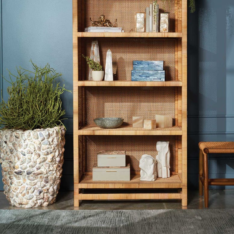 A tall wooden bookshelf with decor and books stands beside the Made Goods Borsala Oyster Shell Planter, Small, filled with greenery, and a woven bench set against a blue wall.