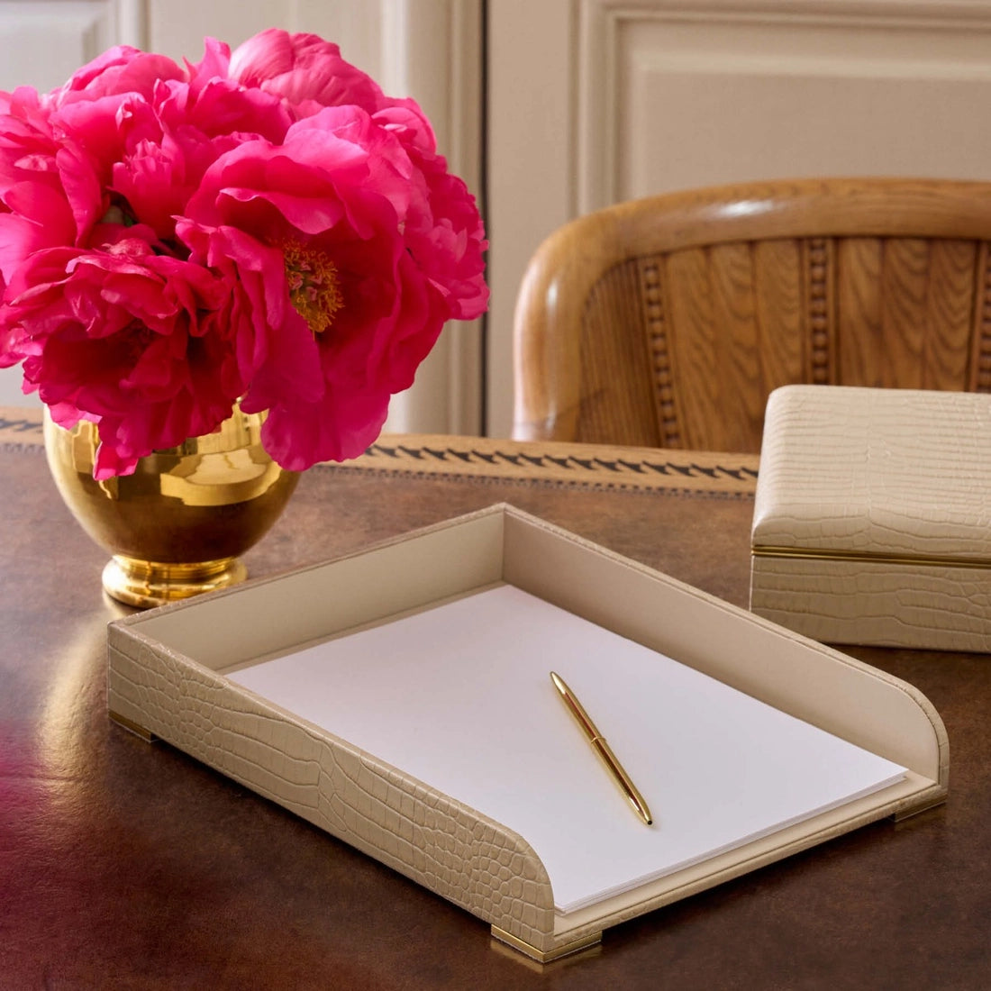 An Aerin Classic Croc Leather Paper Tray in Fawn holds a stack of white paper and a gold pen; next to it on the wooden desk are a beige box and a vase of vibrant pink flowers.