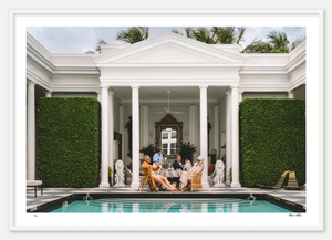 A group of people sit around an outdoor table with drinks at the Nick Mele Fine Art Poker by Nick Mele Photography location, surrounded by classical white columns and neatly trimmed green hedges, next to a rectangular pool with blue water in the foreground.