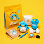Pierre the Penguin Beginner Crochet Kit by The Woobles displayed on a yellow background, featuring yarn, crochet hook, stuffing, plastic needle, eyes, instructions, and small accessories for a complete yarn project.