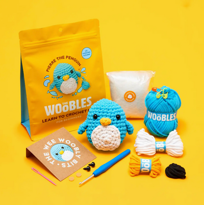 Pierre the Penguin Beginner Crochet Kit by The Woobles displayed on a yellow background, featuring yarn, crochet hook, stuffing, plastic needle, eyes, instructions, and small accessories for a complete yarn project.