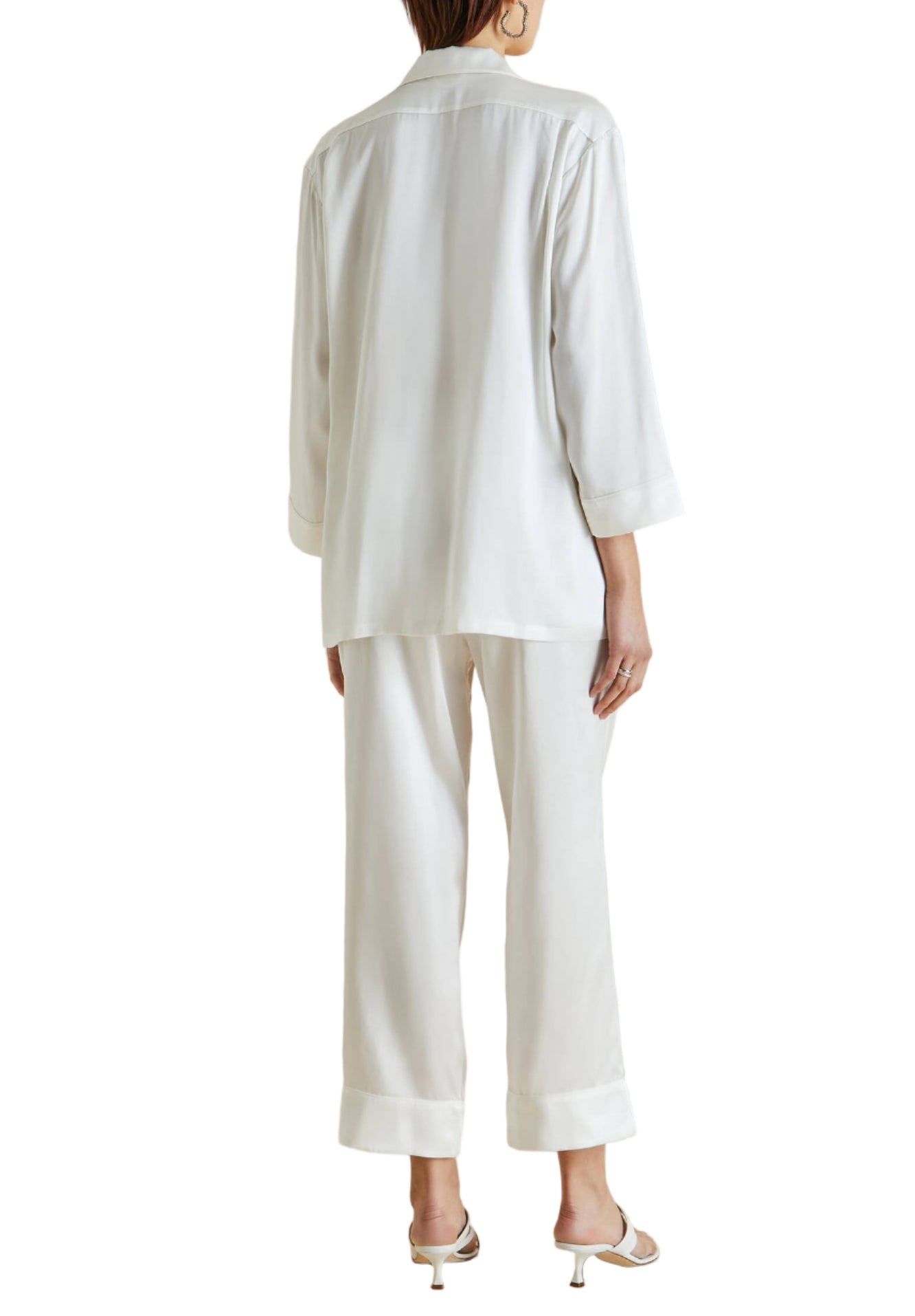 Person wearing Olivia von Halle Fifi Silk Pajamas in white, seen from the back—featuring long sleeves and wide-leg pants, styled with white heeled sandals for an elegant silk pajama look.