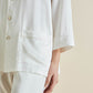 A person wearing Olivia von Halle's Fifi Silk Pajamas—a luxurious white set with a long-sleeve button-up shirt and matching pants—stands with one hand by their side.
