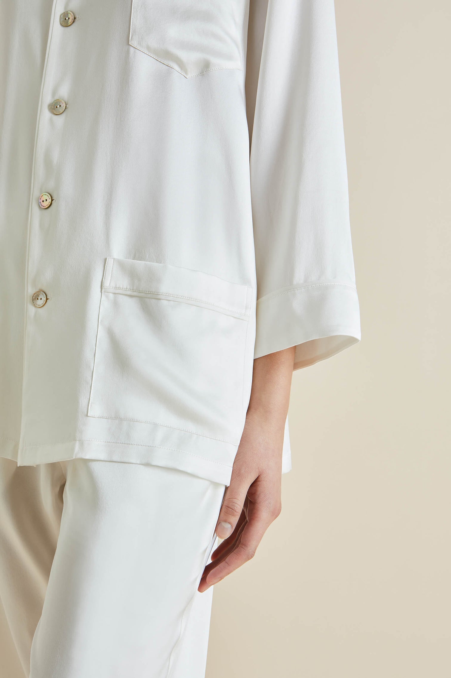 A person wearing Olivia von Halle's Fifi Silk Pajamas—a luxurious white set with a long-sleeve button-up shirt and matching pants—stands with one hand by their side.