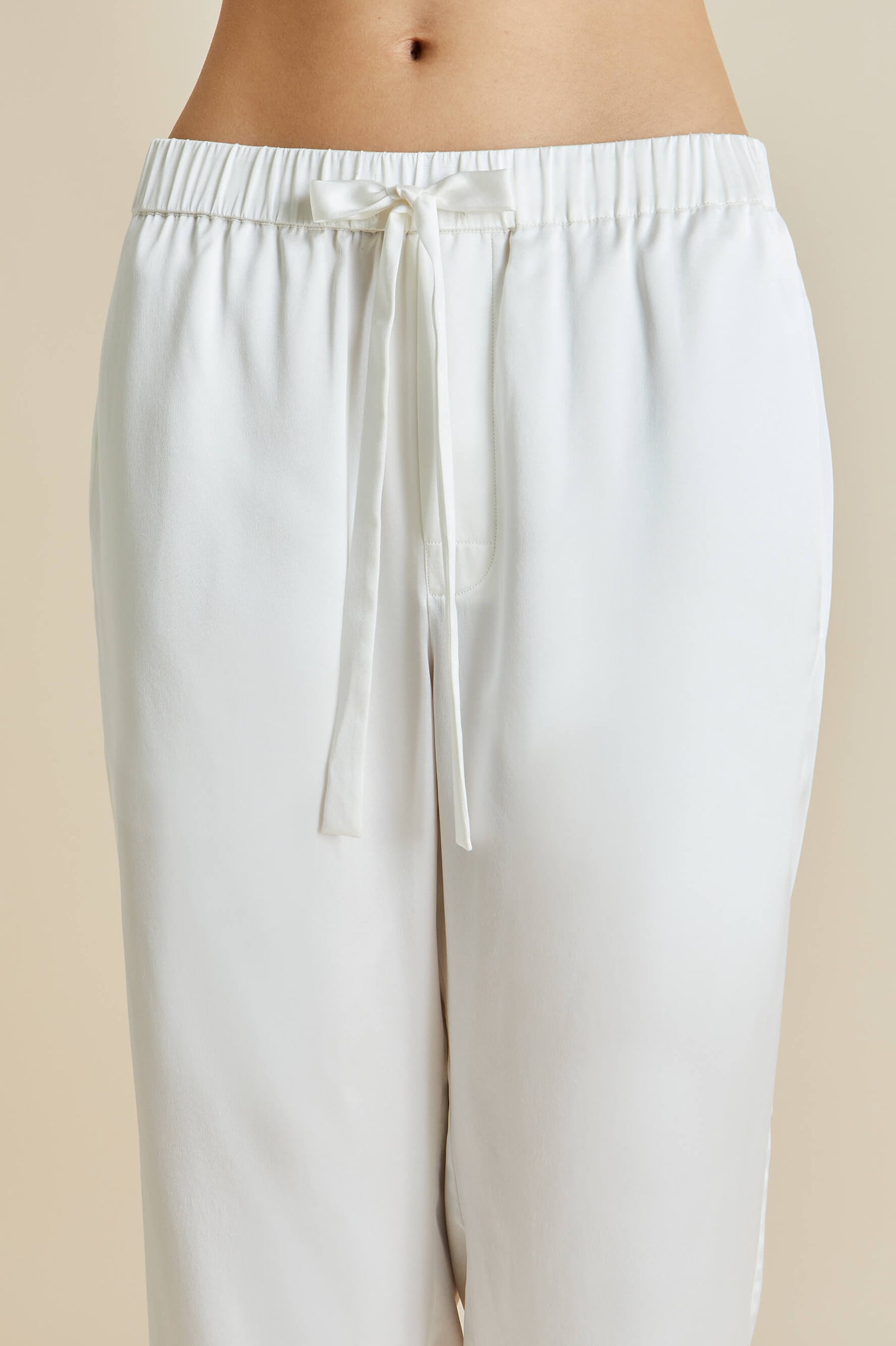Close-up of a person's lower torso wearing loose-fitting white Fifi Silk Pajama pants by Olivia von Halle, featuring an elastic waistband and tied drawstring.