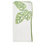 The Kim Seybert Calypso Napkin Set includes 4 white linen napkins, each embroidered with green monstera leaves and finished with a matching green border along one edge.