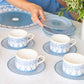 A person arranges a stack of Pink House Mustique Azure Blue Dinnerware Collection plates on a table set with matching teacups, saucers, and a platter. A potted plant is visible to the right.