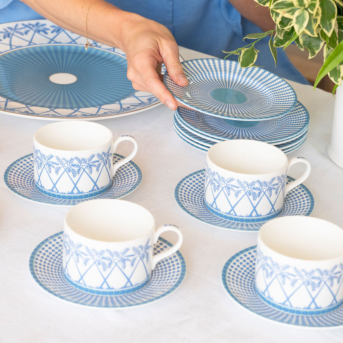 A person arranges a stack of Pink House Mustique Azure Blue Dinnerware Collection plates on a table set with matching teacups, saucers, and a platter. A potted plant is visible to the right.