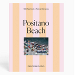 The Piecework Slim Aarons Puzzle Collection features “Positano Beach,” a 1000-piece jigsaw puzzle crafted from recycled paper and depicting a vibrant photograph of a crowded beach with colorful umbrellas and lounge chairs. The puzzle comes packaged in a box decorated with pink and peach stripes.