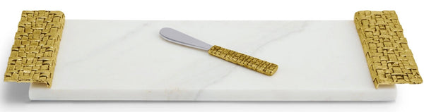 A Michael Aram Palm Cheese Board with Spreader, with a textured antique goldtone handle, lies on a white marble serving board flanked by woven bamboo accents.