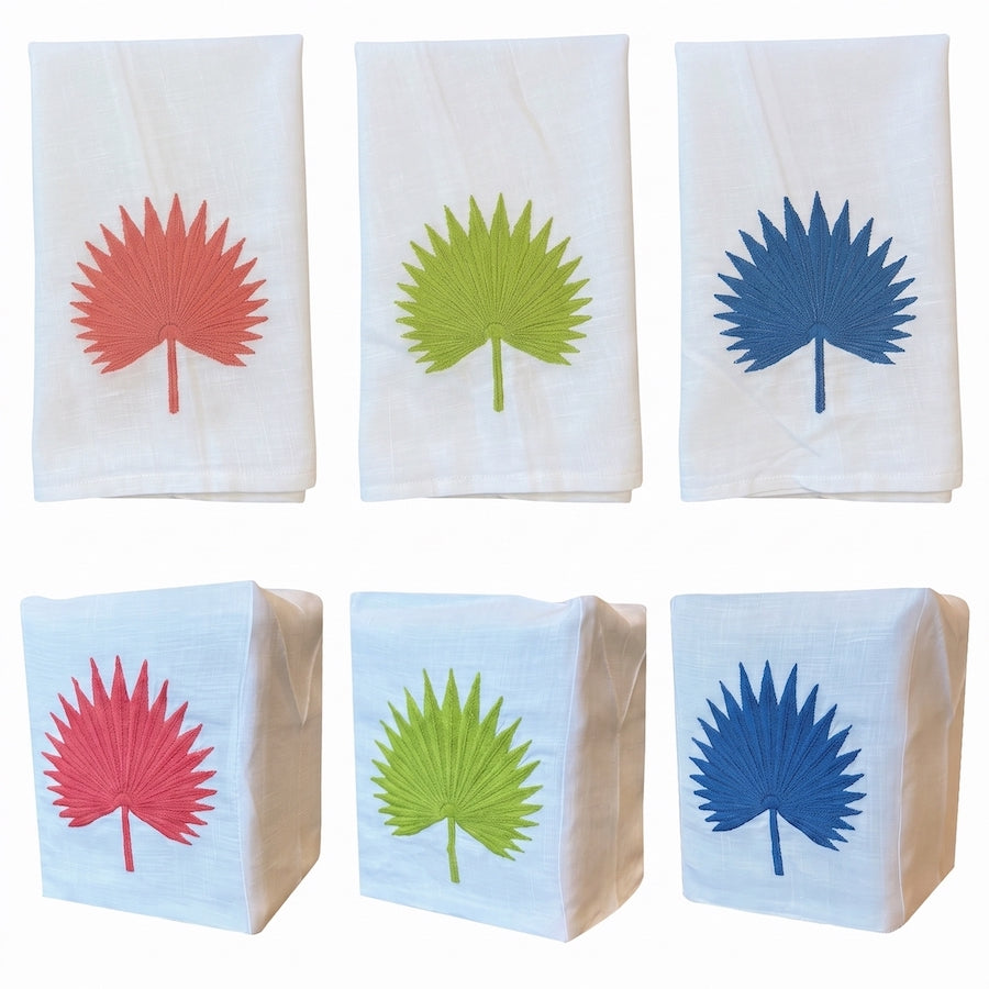 Six white Italian linen tip towels from Haute Home’s Palm Leaf Bath Collection, each featuring an embroidered palm leaf design in red, green, or blue; three towels are laid flat and three are folded into rectangles, all arranged in two rows.