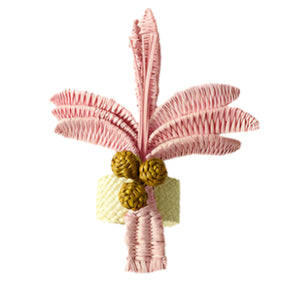 The Duggan Society Palm Napkin Ring is a decorative object made to resemble an Iraca Palm, with handwoven pink fronds, three yellow woven fruits, and a cream-colored cylindrical base.