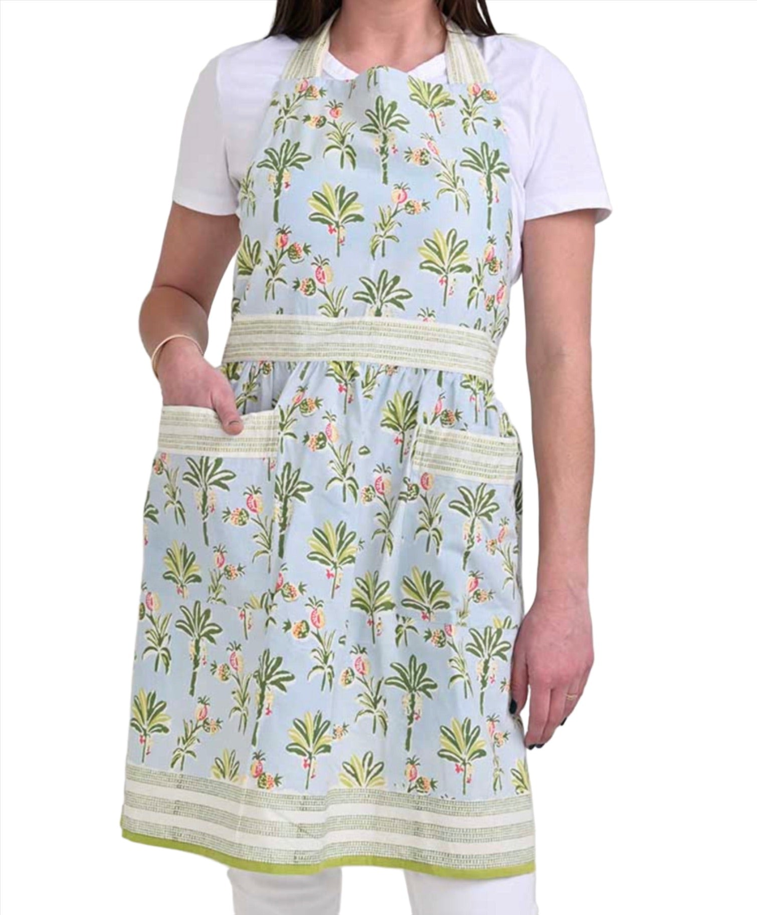 A person wearing a white shirt and a pastel blue, hand block printed cotton apron with green palm tree and floral patterns. The adjustable kitchen apron features two front pockets and a waist tie—an ideal choice for stylish chef gifts from the Pomegranate Inc Apron Collection by Pomegranate Inc.