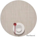 A white plate and bowl with a red spoon sit on a round Chilewich placemat in Parchment, featuring a Mini Basketweave design made from Terrastrand yarns. This phthalate-free placemat ensures safe, stylish dining.