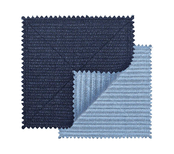 Two square fabric swatches, one dark blue and one light blue with zigzag edges, overlap to display the textured, ribbed basketweave pattern of the Kim Seybert Vista Reversible Placemat.