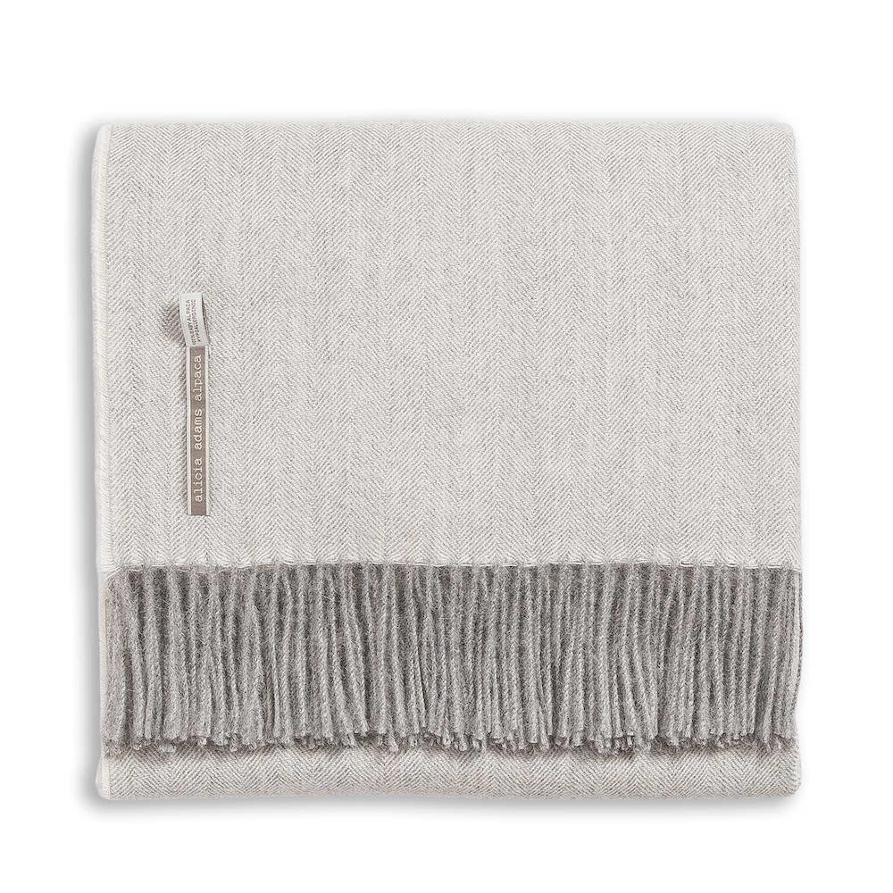 Alicia Adams Alpaca's Classic Herringbone Throw Collection features a folded light gray baby alpaca wool blanket with a darker gray fringed edge and fabric label, ethically crafted in Peru.