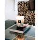 A Baobab Collection Pearls Sapphire Candle in a glass holder sits atop a coffee table book on a reflective glass table, with a patterned chair in the background.