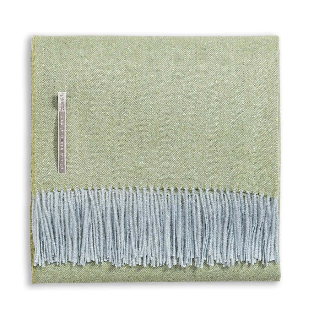 A folded light green Classic Herringbone Throw by Alicia Adams Alpaca, featuring light blue fringes and a fabric label on one side, ethically made in Peru.