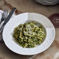 A white bowl of pasta coated in green pesto sauce and topped with grated cheese, inspired by recipes from Via Carota by Penguin Random House, sits on a marble table beside cutlery and stacked plates.