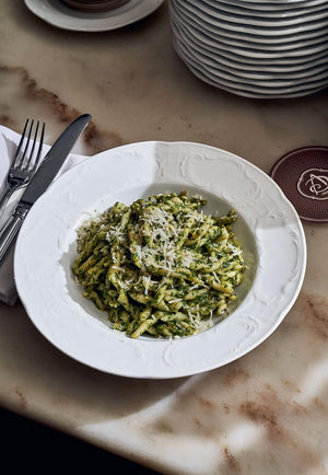 A white bowl of pasta coated in green pesto sauce and topped with grated cheese, inspired by recipes from Via Carota by Penguin Random House, sits on a marble table beside cutlery and stacked plates.