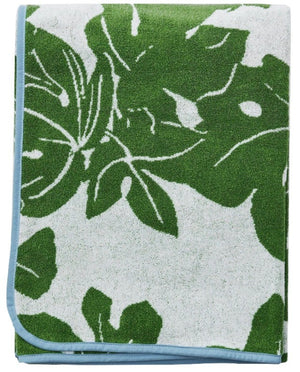 The Weezie x Peter Dunham Fig Beach Towel by Weezie Towels features luxurious folded green fabric with white leaf patterns and a blue border, offering an oversized plush feel ideal for beach lounging.