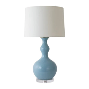 The Pecos Lamp in Nile Blue by Paul Schneider Ceramics features a rounded blue ceramic base and a white cylindrical shade on a clear stand, adding modern elegance and refined lighting to any space.