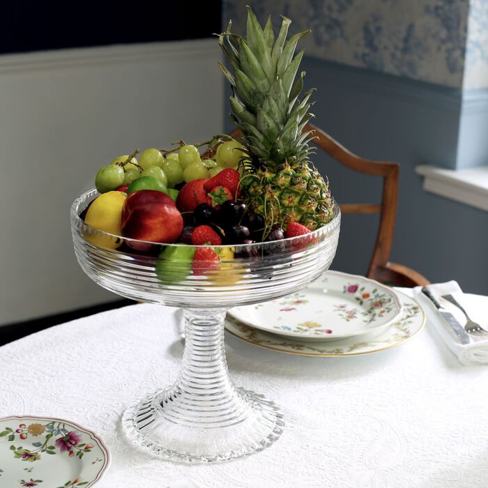 A handmade William Yeoward Crystal Phoebe Centrepiece bowl, brimming with fresh pineapple, grapes, strawberries, and peaches, rests on a white tablecloth beside floral-patterned plates.