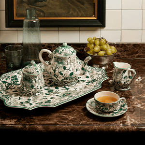 The Pivoines Vertes Dinnerware Collection by Gien France is set on a marble countertop, accompanied by a glass bottle, a bowl of grapes, and a cup of tea.