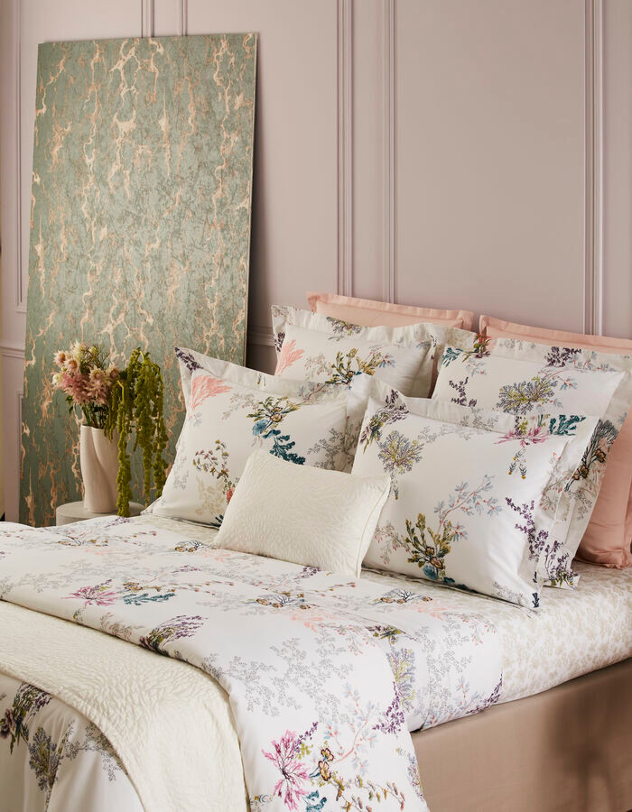 A neatly made bed features Yves Delorme's Naiades Bedding Collection in white floral organic cotton, layered with pillows and a cream blanket, beside a textured green art panel and a vase of greenery in gentle light.