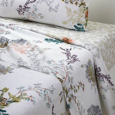 A neatly made bed features Yves Delorme's Naiades Bedding Collection—white sheets and pillowcases with colorful botanical prints, plus an organic cotton reversible duvet cover made in France.