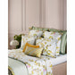 A neatly made bed features Yves Delorme's Luciole Bedding Collection, showcasing floral-patterned sheets and a reversible organic cotton sateen duvet cover, accented by green, white, and yellow pillows against a beige paneled wall.