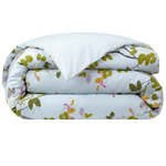 A folded comforter from the Yves Delorme Luciole Bedding Collection features a light blue background with green and pink floral branches, crafted from organic cotton sateen. Pair with its reversible duvet cover for versatile style.