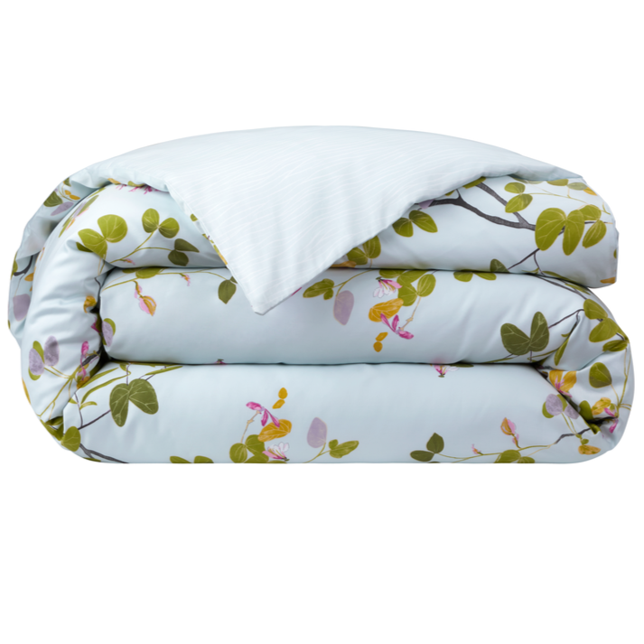 A folded comforter from the Yves Delorme Luciole Bedding Collection features a light blue background with green and pink floral branches, crafted from organic cotton sateen. Pair with its reversible duvet cover for versatile style.