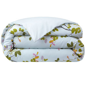 A folded comforter from the Yves Delorme Luciole Bedding Collection features a light blue background with green and pink floral branches, crafted from organic cotton sateen. Pair with its reversible duvet cover for versatile style.