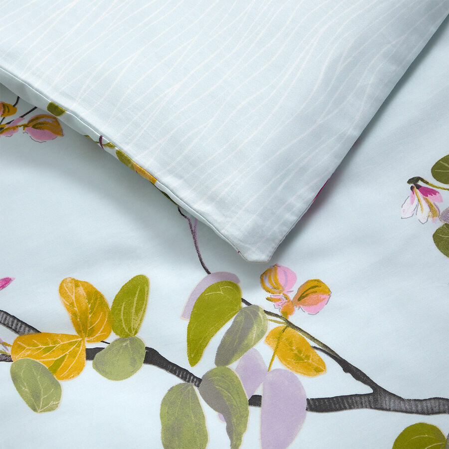 Close-up of a pillow and bedsheet from the Yves Delorme Luciole Bedding Collection, crafted in organic cotton sateen, featuring a light blue background with floral branch patterns in green, yellow, and pink.