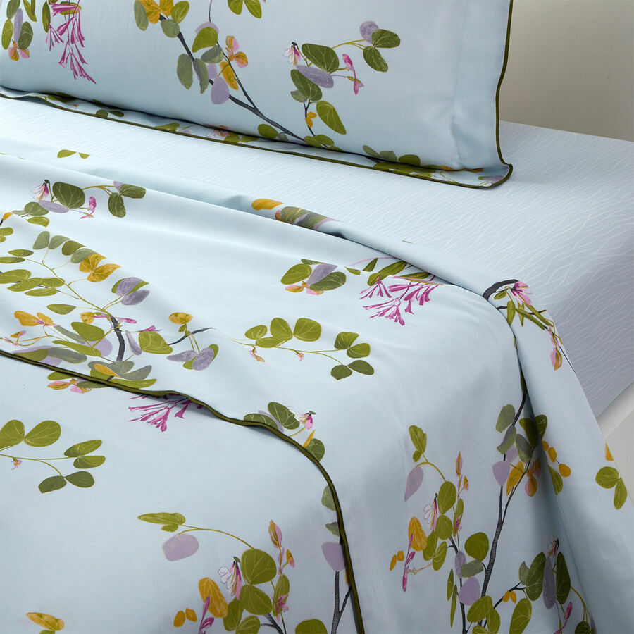Light blue bedding set from the Yves Delorme Luciole Bedding Collection, featuring a green leaf and pink flower print on organic cotton sateen, includes pillowcases and a reversible duvet cover neatly spread on the bed.