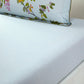 The Luciole Bedding Collection by Yves Delorme features a light blue organic cotton sateen fitted sheet with a subtle wavy texture, paired with a pillowcase adorned with green leaves and pink flowers.