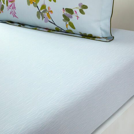 The Luciole Bedding Collection by Yves Delorme features a light blue organic cotton sateen fitted sheet with a subtle wavy texture, paired with a pillowcase adorned with green leaves and pink flowers.