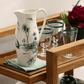 A ceramic pitcher from the Depareillees Vert Dinnerware and Serveware Collection by Gien France sits on a folded cloth next to four striped glasses on a wooden serving cart—ideal for effortless table decor.