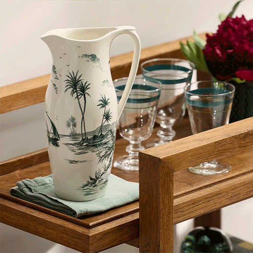 A ceramic pitcher from the Depareillees Vert Dinnerware and Serveware Collection by Gien France sits on a folded cloth next to four striped glasses on a wooden serving cart—ideal for effortless table decor.