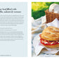 The Picnic round loaf from Simon & Schuster, filled with mozzarella, salami, and tomato, sits invitingly on a plate next to a knife and picnic basket on lush green grass—making it a perfect choice for summer picnics and al fresco dining.