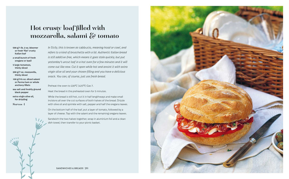 The Picnic round loaf from Simon & Schuster, filled with mozzarella, salami, and tomato, sits invitingly on a plate next to a knife and picnic basket on lush green grass—making it a perfect choice for summer picnics and al fresco dining.